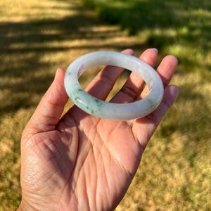 May include: A smooth, circular bangle bracelet crafted from translucent, pale white stone with distinct, mottled patches of vibrant green color throughout the band. The jewelry piece features a polished, rounded surface and a consistent thickness.