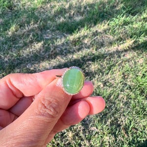 May include: A gold ring featuring a large, oval-shaped, light green gemstone. The gemstone is surrounded by a halo of small, clear stones. The ring is held against a backdrop of green grass.