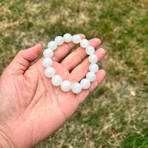 May include: A white beaded bracelet with large, round beads. The bracelet is held in a hand against a backdrop of green grass. The beads have a smooth, polished appearance and are strung together to form a circular shape.