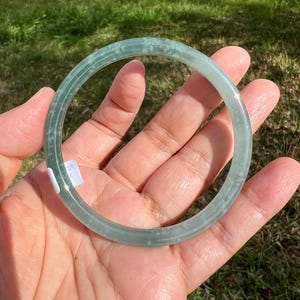 May include: A pale green jade bangle bracelet. The circular bracelet is held in a hand, highlighting its smooth, polished surface. The jade has subtle colour variations, with a slightly textured appearance. A small price tag is visible.