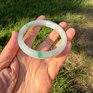 May include: A smooth, circular jade bangle bracelet featuring a translucent pale green base with a distinct, vibrant emerald green spot. A small white rectangular sticker is attached to the surface, displaying the printed text WP22609 100360.