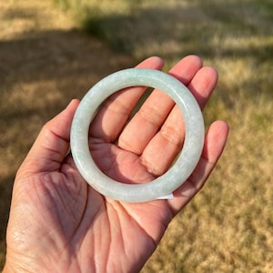 May include: A smooth, circular bangle bracelet crafted from pale, translucent green jade stone. The bangle features a polished, rounded surface with subtle variations in color and texture throughout the material, creating a soft, cloudy appearance.