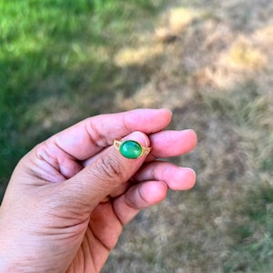 Op de afbeelding: Een gouden ring met een gladde, ovaalvormige groene edelsteen in het midden. De band van de ring heeft een delicaat, opengewerkt ontwerp aan de zijkanten, wat een gedetailleerd beeld geeft van het metaalwerk rondom de levendige groene steen.