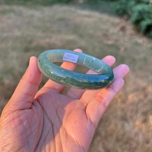 Puede incluir: Un brazalete de jade pulido y liso que presenta un color verde translúcido con inclusiones moteadas más oscuras. El interior de la banda circular muestra una pequeña etiqueta rectangular blanca con el texto impreso WP212608 100827.