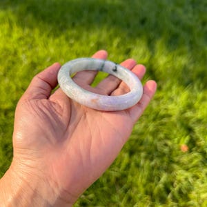 May include: A smooth, rounded bangle bracelet crafted from polished stone. The bangle features a translucent appearance with a blend of pale lavender, soft white, and deep green mottled inclusions throughout the circular band structure.