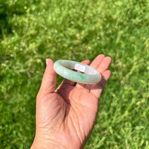Può includere: Un bracciale rigido in giada liscio e circolare, caratterizzato da un colore verde pallido traslucido con una piccola inclusione verde smeraldo brillante. Un piccolo adesivo rettangolare bianco con testo nero è applicato all'interno.