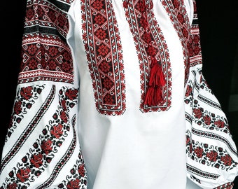 Puede incluir: Una blusa blanca de manga larga con intrincados bordados geométricos y florales en rojo y negro en el pecho y las mangas. La prenda incluye un cuello redondo con un cierre de cordón terminado con dos borlas rojas en el centro.