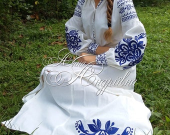 May include: A white embroidered dress with blue floral patterns. The dress has long sleeves and a flowing skirt. The model is wearing the dress and sitting in a grassy area.