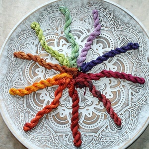 Op de afbeelding: Een verzameling kleurrijk borduurgaren, gerangschikt in een cirkelvormig patroon op een decoratief, wit houten bord. De garenkleuren variëren van warme oranje- en roodtinten tot koele groen- en paars tinten voor knutselprojecten.