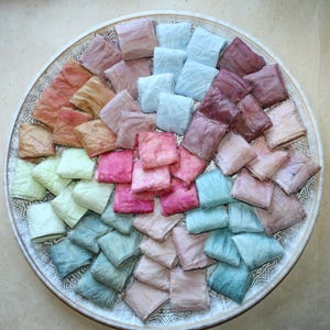 Op de afbeelding: Een ronde decoratieve schaal bevat een verzameling gevouwen stoffen linten in een kleurenspectrum met tinten blauw, groen, roze, paars en oranje. De linten zijn in een cirkelvormig patroon gerangschikt en tonen verschillende texturen.