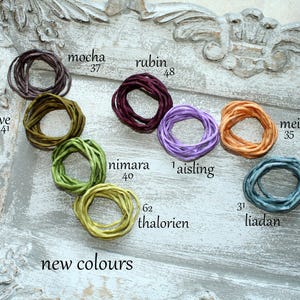 Puede incluir: Ocho cordones enrollados de varios colores etiquetados con nombres y números: mocha 37, olive 41, nimara 40, thalorien 62, rubin 48, aisling 1, meilin 35 y liadan 31. El texto new colours está impreso en la parte inferior izquierda.