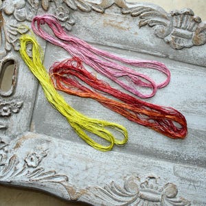 May include: Three hanks of embroidery thread in bright yellow, variegated pink, and variegated red-orange. The threads are loosely coiled and displayed on a weathered, gray, rectangular decorative tray with ornate carved floral edges.