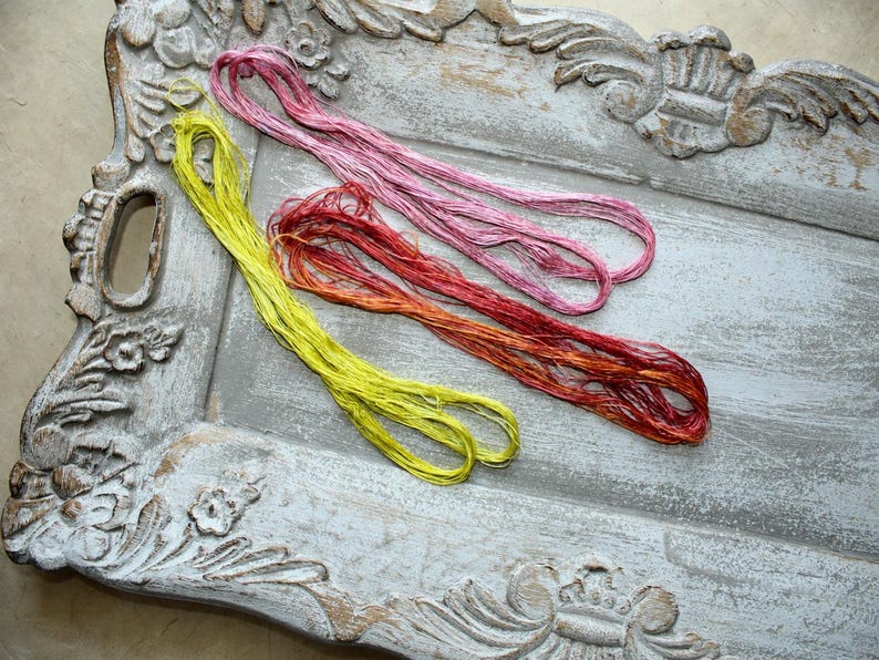 May include: Three hanks of embroidery thread in bright yellow, variegated pink, and variegated red-orange. The threads are loosely coiled and displayed on a weathered, gray, rectangular decorative tray with ornate carved floral edges.