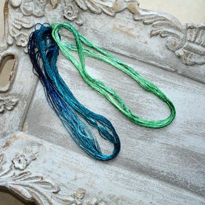 May include: Two long, looped bundles of embroidery thread. One bundle features a gradient of deep navy blue to bright turquoise, while the other bundle consists of a vibrant, solid lime green. The threads have a smooth, lustrous, and silky texture.