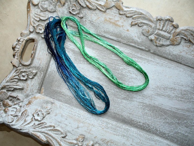 May include: Two long, looped bundles of embroidery thread. One bundle features a gradient of deep navy blue to bright turquoise, while the other bundle consists of a vibrant, solid lime green. The threads have a smooth, lustrous, and silky texture.