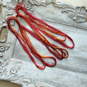 May include: A bundle of variegated embroidery thread featuring a blend of deep red, crimson, and golden orange hues. The thread is loosely coiled and consists of multiple thin strands with a slight sheen, showcasing a gradient of warm autumn colors.