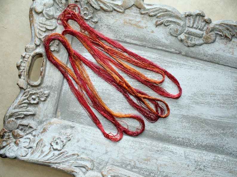 May include: A bundle of variegated embroidery thread featuring a blend of deep red, crimson, and golden orange hues. The thread is loosely coiled and consists of multiple thin strands with a slight sheen, showcasing a gradient of warm autumn colors.