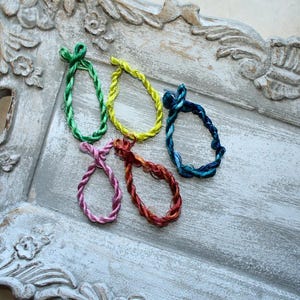 May include: Five circular hair ties made of twisted, shiny cord in vibrant colors including green, yellow, dark blue, pink, and reddish-brown. Each hair tie features a small, knotted loop at the top, showcasing a smooth, reflective texture.