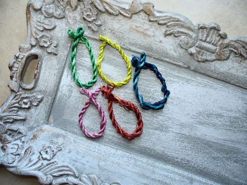 May include: Five circular hair ties made of twisted, shiny cord in vibrant colors including green, yellow, dark blue, pink, and reddish-brown. Each hair tie features a small, knotted loop at the top, showcasing a smooth, reflective texture.