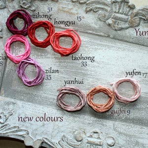 May include: A collection of nine small, circular coils of colored ribbon or cord, labeled with names and numbers including zihong 51, hongyu 15, fenxia 8, taohong 33, zilan 35, yanhui, yufen 17, guifei 9, and Yunjin, arranged on a distressed gray tray.