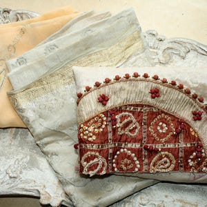 Könnte beinhalten: Eine Sammlung gefalteter Stoffe, darunter ein pfirsichfarbenes Textil mit Silberstickerei, ein hellgrauer Stoff mit metallischem Goldrand und ein weißer Stoff mit dunkelrotem Karomuster, Perlenstickerei und metallischen Goldfäden.