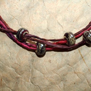 May include: A necklace featuring multiple strands of twisted magenta and deep red silk ribbon. Four smooth, round, dark-toned beads with speckled patterns are threaded onto the ribbons, spaced out along the length of the necklace.