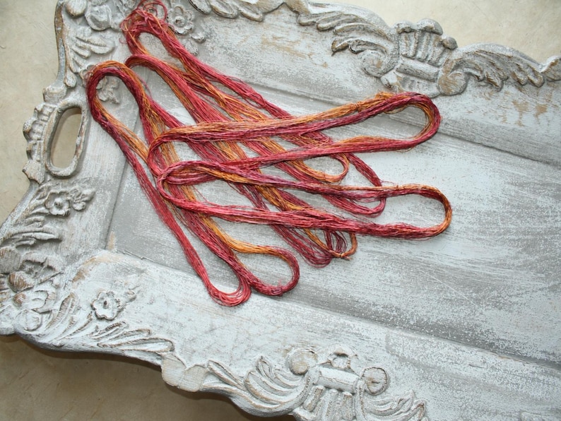 May include: A bundle of variegated embroidery thread in shades of deep red, orange, and gold, coiled in loose loops. The thread features a textured, slightly metallic appearance with a multi-tonal color gradient throughout the strands.