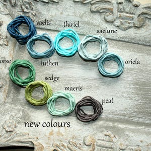 Pode incluir: Uma coleção de dez elásticos de cabelo circulares de tecido em vários tons de azul, verde e castanho, rotulados com o texto vaelis, thiriel, saelune, gorse, luthen, oriela, sedge, maeris, peat e new colours, dispostos numa bandeja cinzenta.