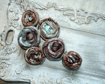 May include: Six brown and teal fabric rosettes arranged in a semi-circle on a white, ornate tray.