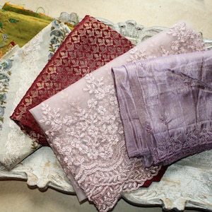 Op de afbeelding: Een assortiment gevouwen stoffen in verschillende kleuren en texturen. De stoffen omvatten een groen geborduurd stuk, een wit en blauw patroon, een bordeauxkleurige brokaat, een roze kant en een lavendelkleurige zijde. De stoffen zijn gerangschikt op een decoratief dienblad.