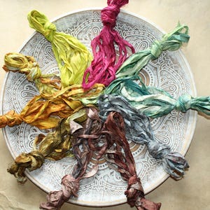 Puede incluir: Una colección de cintas de seda teñidas a mano dispuestas en un patrón circular sobre un plato decorativo. Las cintas presentan una variedad de colores que incluyen verde lima, magenta, verde azulado, gris, marrón, amarillo mostaza y verde oliva.