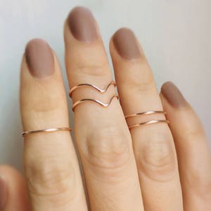 May include: A set of four gold-toned rings, including a double-band ring and a V-shaped ring, worn on a hand with nude nail polish.