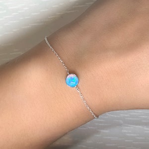 Könnte beinhalten: Ein zartes Silberarmband mit einem runden, irisierenden blauen Opal-Edelstein. Der Opal ist in einer einfachen Lünettenfassung gefasst und mit einer feinen Kette verbunden. Das Armband ist für das Handgelenk konzipiert.