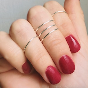 May include: A hand wearing multiple thin silver rings on the index finger and a red nail polish on the ring finger, middle finger, and pinky finger.