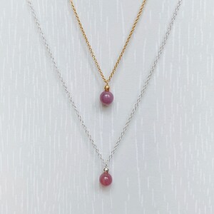 May include: Two delicate necklaces with a single, round, purple gemstone pendant on each. The top necklace has a gold chain and the bottom necklace has a silver chain.