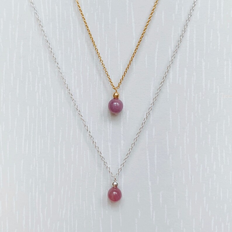 May include: Two delicate necklaces with a single, round, purple gemstone pendant on each. The top necklace has a gold chain and the bottom necklace has a silver chain.