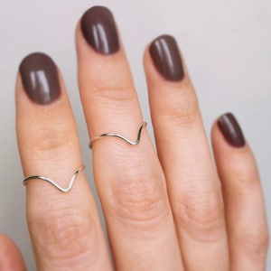 May include: Two thin, polished silver-coloured rings, each featuring a simple V-shaped chevron design. The rings are worn on the fingers, showcasing a smooth, metallic finish and a minimalist, pointed silhouette that sits flat against the skin.