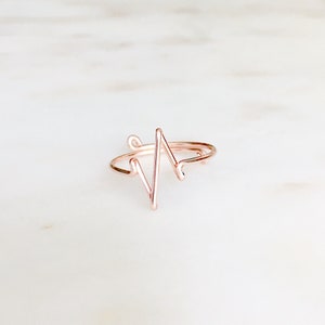 May include: A rose gold-colored wire ring featuring a continuous, angular heartbeat or EKG line design. The thin, metallic band is shaped into a sharp, zigzagging pattern that rises from the center of the ring, creating a distinct, open-loop silhouette.