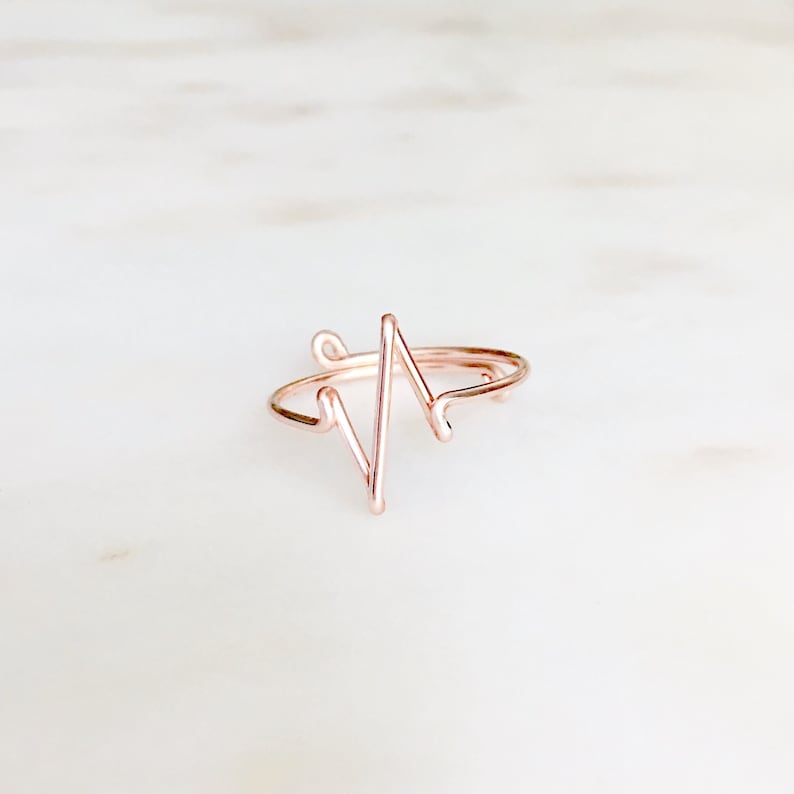 May include: A rose gold-colored wire ring featuring a continuous, angular heartbeat or EKG line design. The thin, metallic band is shaped into a sharp, zigzagging pattern that rises from the center of the ring, creating a distinct, open-loop silhouette.