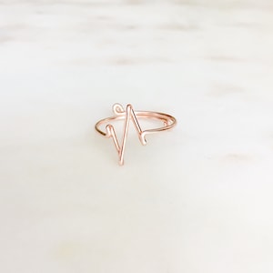 May include: A rose gold-colored wire ring featuring a continuous, angular heartbeat EKG line design. The thin, metallic band is formed into a singular, sharp zigzag pattern that rises from the circular base, creating a distinct, minimalist, and open-ended shape.