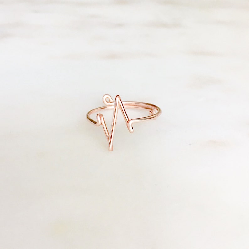 May include: A rose gold-colored wire ring featuring a continuous, angular heartbeat EKG line design. The thin, metallic band is formed into a singular, sharp zigzag pattern that rises from the circular base, creating a distinct, minimalist, and open-ended shape.