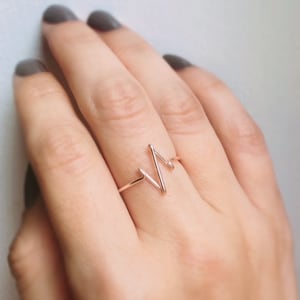 May include: A rose gold ring featuring a thin, continuous wire band shaped into a stylized heartbeat or EKG line pattern. The metal has a smooth, polished finish, and the design consists of a single sharp peak and a smaller secondary wave.