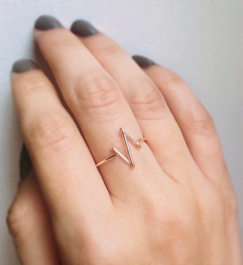 May include: A rose gold ring featuring a thin, continuous wire band shaped into a stylized heartbeat or EKG line pattern. The metal has a smooth, polished finish, and the design consists of a single sharp peak and a smaller secondary wave.