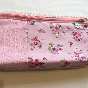 May include: A pink zippered pouch with a floral print and a small flower charm. The pouch has a separate pocket on the front with a white fleur-de-lis pattern.