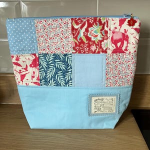 May include: A patchwork fabric pouch with a light blue base and a patterned top. The top features squares of fabric with floral, polka dot, and elephant designs. A small label is sewn on the front.