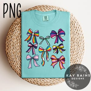 May include: A turquoise blue t-shirt with a design of colorful bows. The bows are in various colors and patterns, including rainbow, pink, blue, and purple. The text "KAY RAINS DESIGNS" is printed on the bottom right corner of the shirt.