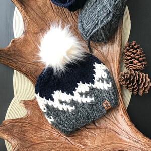 Puede incluir: Un gorro de punto con un pomp&oacute;n blanco, que presenta un patr&oacute;n de monta&ntilde;a azul y gris. El gorro tiene una etiqueta de cuero con las iniciales "MK".