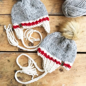 Puede incluir: Dos gorros de punto grises con ribetes blancos y rojos y pompones blancos. Los gorros tienen lazos blancos con borlas.