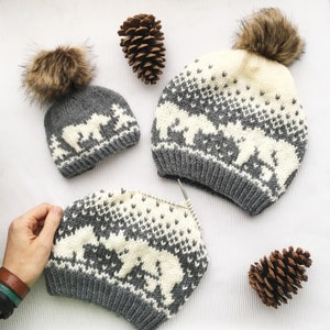May include: Three knitted hats with white polar bear designs on a gray and white background. The hats have fluffy pom poms on top. The hats are on a white surface with pine cones.