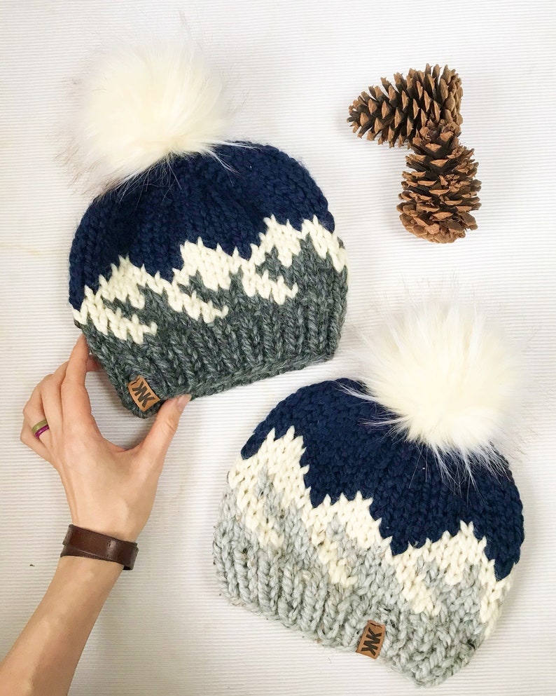 Puede incluir: Dos gorros de invierno de punto con pompones blancos. Los gorros son azul marino, gris y blanco con un patr&oacute;n geom&eacute;trico. Los gorros tienen una etiqueta de cuero marr&oacute;n con las letras "MK" en ellos.